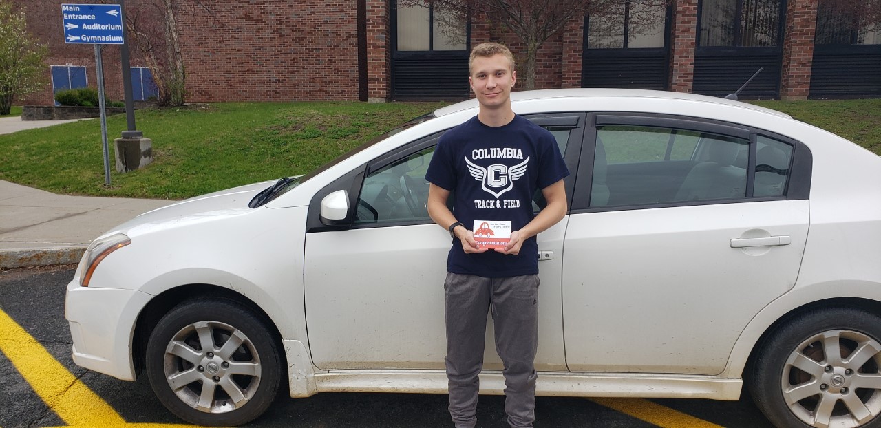A young man posing with a white car, holding up a card that says "Congratulations!"