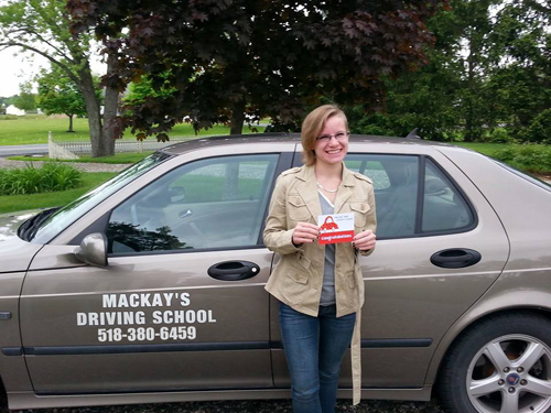 A woman posing with a silver car. A decal on the side of the car reads: Mackay's Driving School.