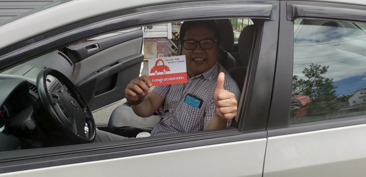 A young driver in a car, giving a "thumbs up" to the camera and holding up a red card that says "Congratulations!"